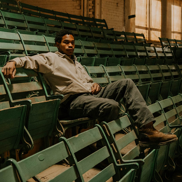 A man sits in the stands at the cattle sale barn, posing and wearing workwear.